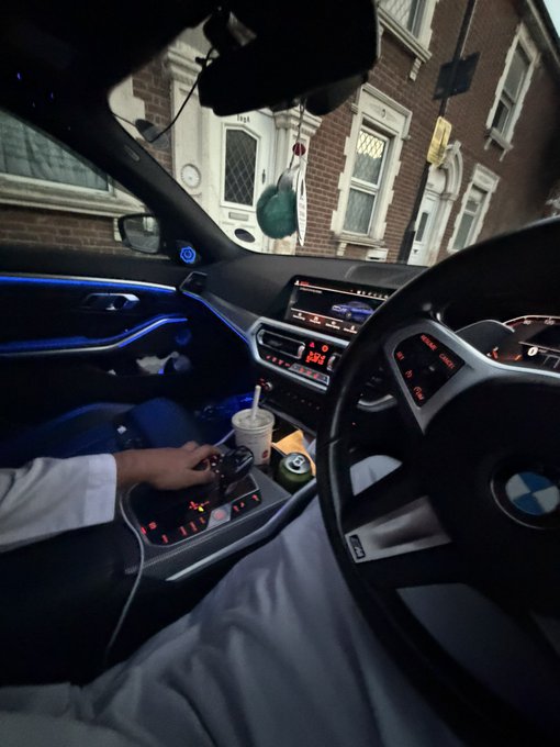 A passenger's view from inside a BMW (logo visible on the steering wheel) shows the driver reaching toward the center console where a McDonald's cup and small snack items sit. The post's reference to "GM bros" contrasts the luxury German vehicle with the everyday American fast-food meal inside, highlighting a humorous clash between high-end and casual.