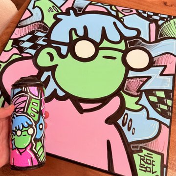 A hand holds a cylindrical container featuring an artistic character design next to a matching painting of the same figure on a table.