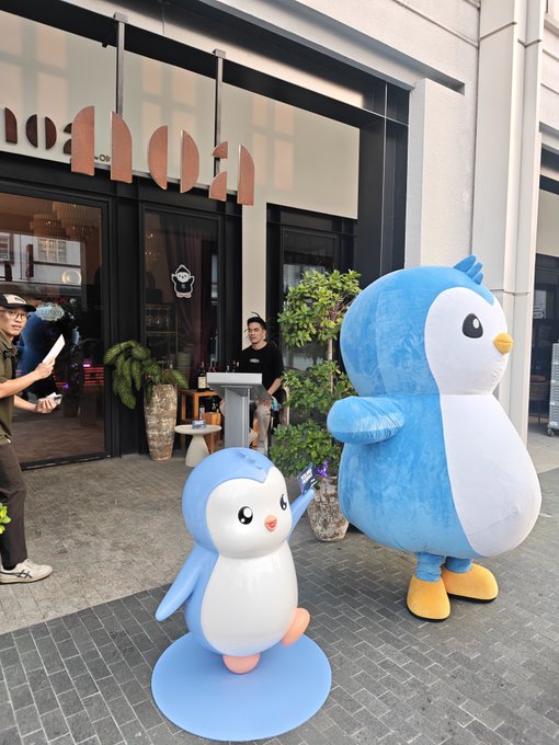 Two penguin mascots (one large blue and one smaller statue) standing outside a storefront, with people visible in the background near the glass windows.