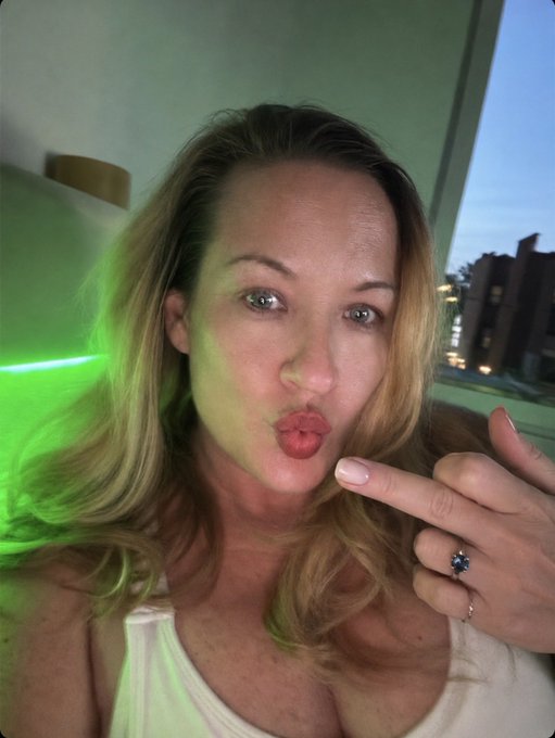 A blonde woman making a kissing face at the camera while displaying a ring on her finger, with a green light illuminating her hair.