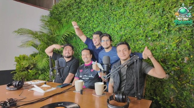 A group of four friends pose cheerfully behind podcast microphones and headphones at a recording session for "No Me Tokens," with the show's logo visible in the upper right corner. The image captures the podcast's second episode, where the Spanish-speaking team discussed crypto gaming concepts including ownership and accessibility for a Latin American audience.