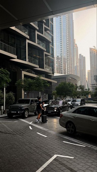 A cityscape view from beneath a building overhang shows a woman with luggage crossing a street with a Jeep and other cars, surrounded by modern skyscrapers.