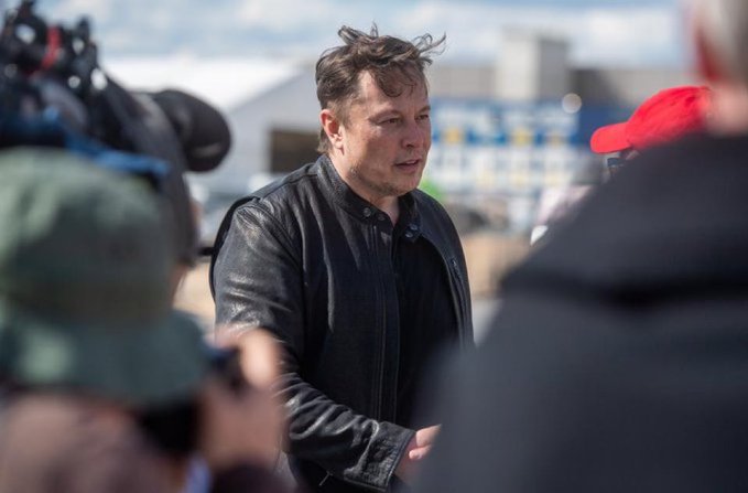 Elon Musk in a black leather jacket walking outdoors while surrounded by people, including a photographer with a camera in the foreground.