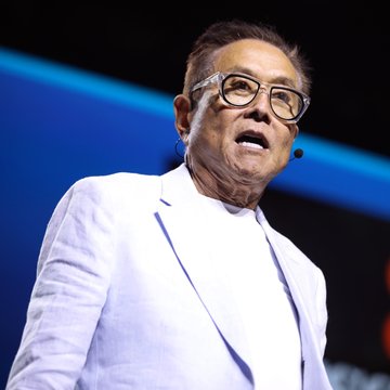 A man wearing glasses and a white suit jacket performs on stage with his mouth open, wearing a small earpiece microphone.