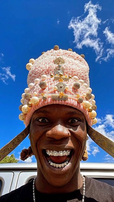 A low-angle portrait of a man smiling widely, wearing a pink knit hat adorned with pearls and gold decorations, displaying diamond-encrusted grillz on his teeth, set against a blue sky.