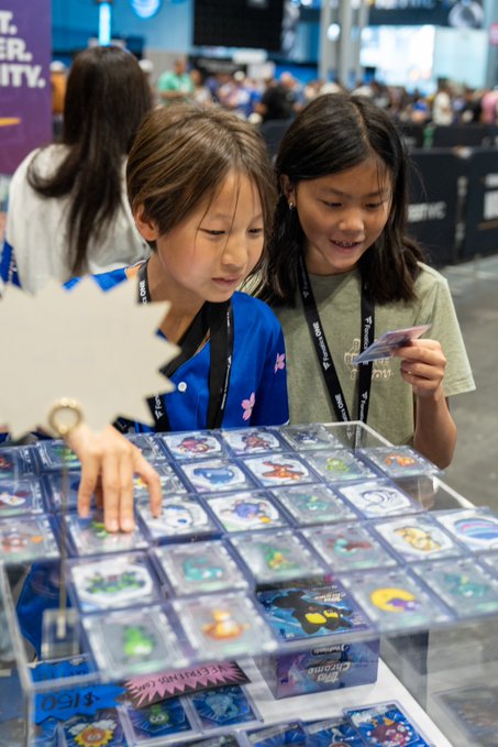 Two young girls browse Pokemon trading cards and merchandise at what appears to be a convention booth, with one girl wearing a blue jacket and reaching toward the card display.