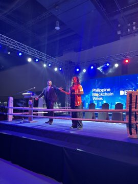 The image shows a stage with two men standing behind a podium, one wearing a suit and the other an orange shirt. They are surrounded by several chairs arranged around the stage, suggesting that they might be participating in a panel discussion or event. The background features a large screen displaying "Philippine Business Chain Week" along with some text, indicating the context of the scene.