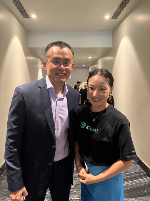 A man in a suit stands next to a woman wearing a black 'X-Hunt' t-shirt and a blue apron in a hallway.