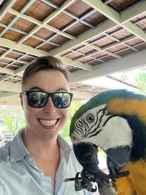 A woman wearing sunglasses smiles for a selfie alongside a blue and yellow macaw perched on her hand.