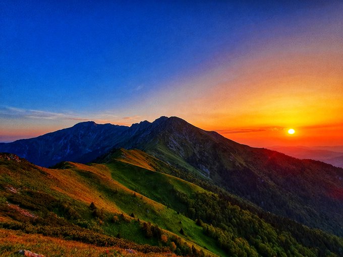A scenic landscape photograph of green mountain ridges under a dramatic sunset sky with the sun glowing orange near the horizon.