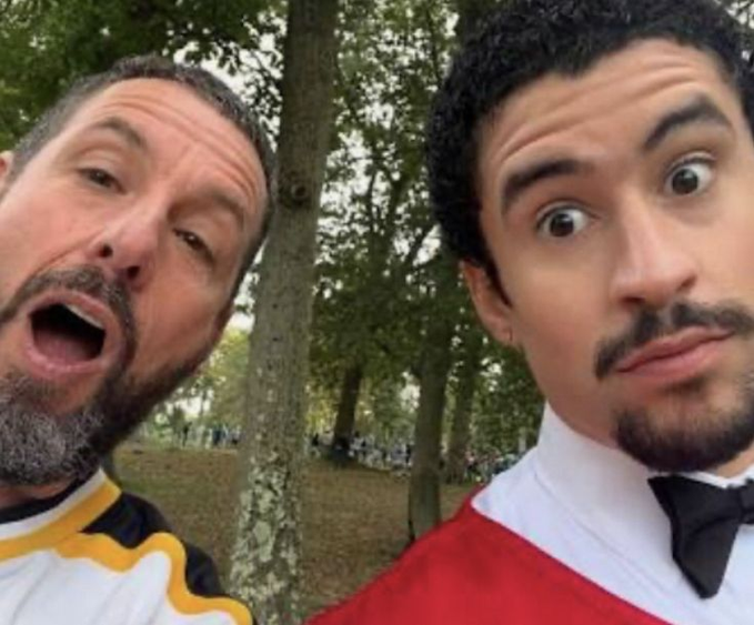 Two men take a close-up selfie outdoors in a wooded area, with the older bearded man on the left making an exaggerated open-mouthed expression while the dark-haired man on the right makes wide-eyed faces.