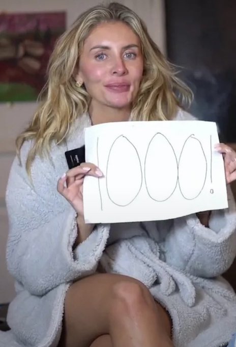 A blonde woman in a fluffy white robe holds up a white card with the number '1000.' written on it in large handwritten digits.