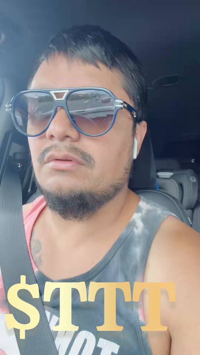 The image features a man wearing sunglasses, a tank top with the text "STTTT", and a beard. He appears to be sitting inside a car, possibly driving or waiting for someone else to join him. The photo has been edited to include the caption "Cooking" in the bottom right corner of the frame.