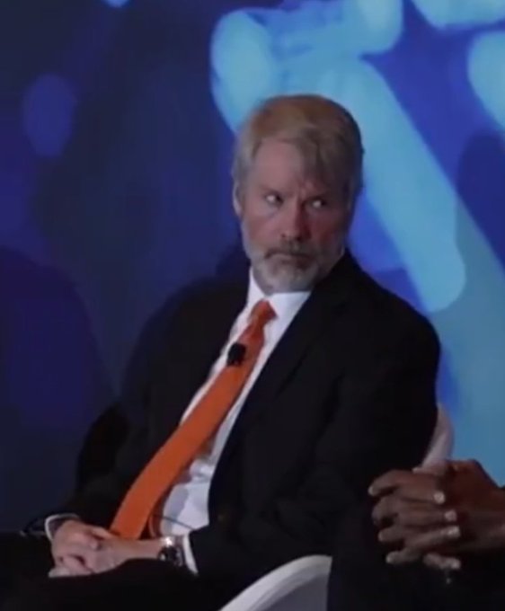 A photo of Julian Assange seated on stage, wearing a black suit with an orange tie, looking to the side with a serious expression.