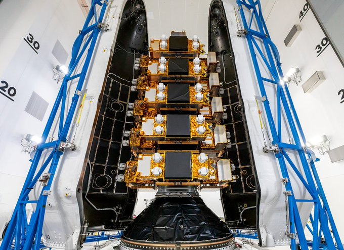 An overhead view shows the interior of a satellite or spacecraft payload fairing in a clean room facility, revealing stacked gold-foiled electronic components and equipment secured between black structural halves, surrounded by blue scaffolding marked with numbers '20' and '30'.