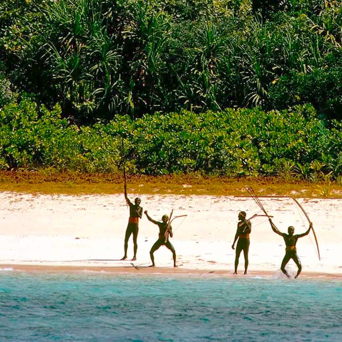 Four indigenous people stand on a white sand beach holding bows, posed against a backdrop of dense green tropical vegetation and turquoise water.