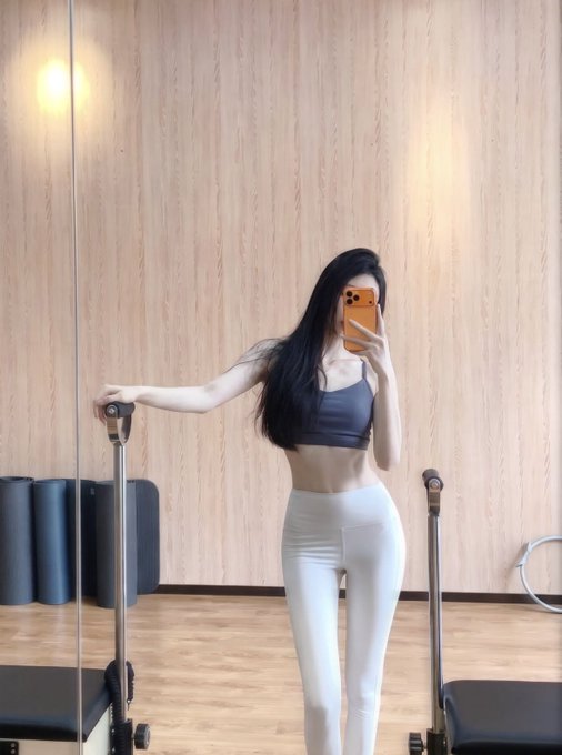 A woman takes a mirror selfie in a pilates studio, wearing a gray sports bra and white leggings while posing next to a reformer machine. The caption's juxtaposition of fitness imagery with a declaration of unwavering commitment to holding Bitcoin creates an ironic contrast between physical discipline and crypto conviction.