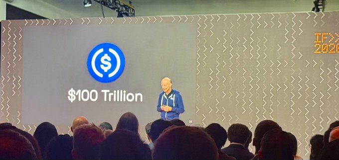 Conference scene with a speaker on stage presenting a slide displaying the USDC logo and '$100 Trillion' to an audience.