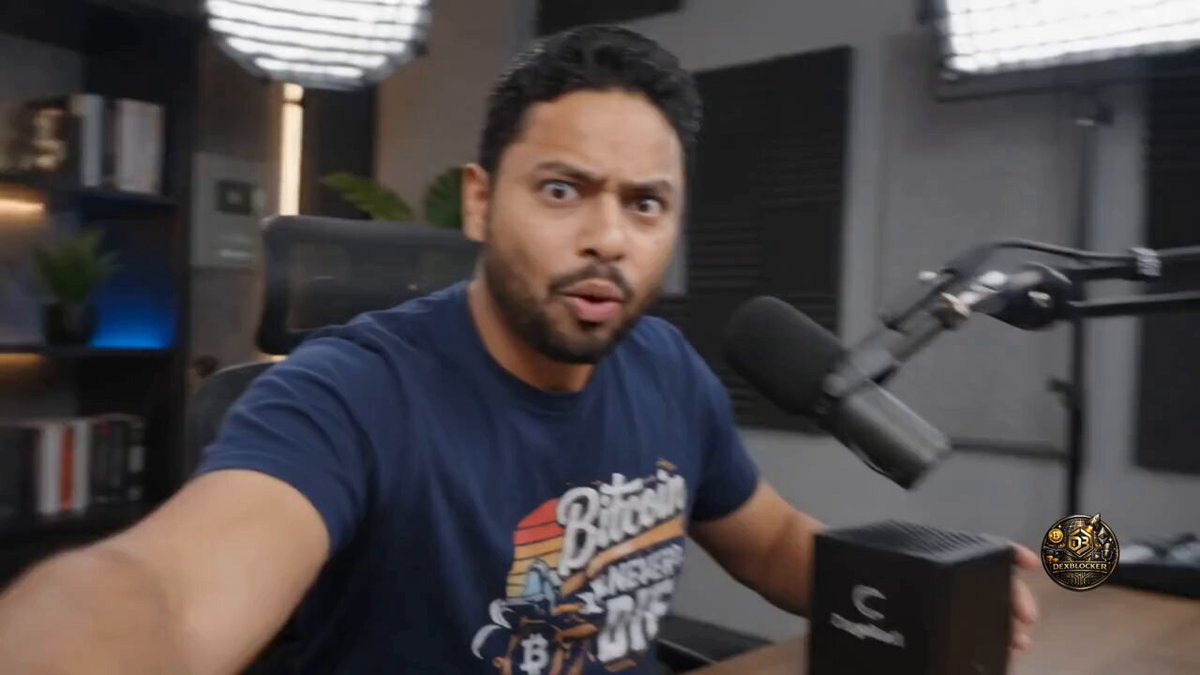 The image shows a man wearing a blue shirt sitting at a desk with a microphone, holding a small cube that appears to be a 100mm power supply unit (PSU). The man seems surprised or intrigued by something happening behind him. The background of the image features a bookshelf filled with various items and a potted plant.