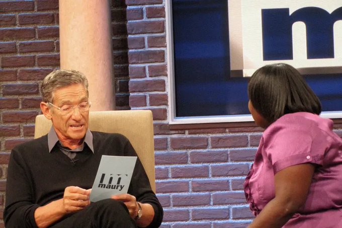 An older man wearing glasses and a dark sweater holds a 'maury' cue card while seated across from a woman in a pink blouse on the set of the Maury show, with a brick wall backdrop visible behind them.