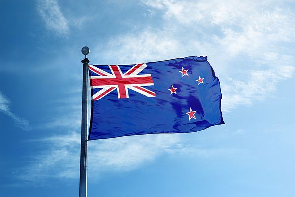 Photograph of the New Zealand flag (blue ensign with Union Jack and red stars) waving on a flagpole against a partly cloudy sky.