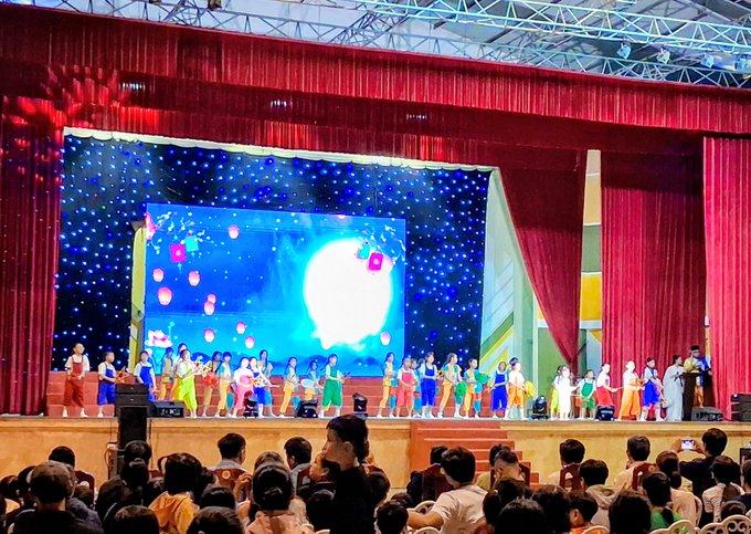 A school stage performance features a large group of children in traditional and colorful costumes performing in front of a large LED screen displaying a moon and lanterns, with red curtains framing the stage and an audience watching.
