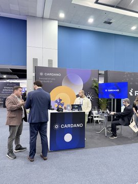 Photo of a Cardano booth at a conference or trade show, with several people standing around the display tables featuring the Cardano logo.