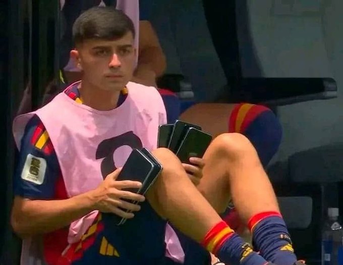 A young Spanish national team player wearing a pink training bib sits on the bench holding multiple smartphones, appearing to multitask between devices. The image humorously illustrates the post's theme of stacking multiple prediction market bets across different platforms like Polymarket, as the player juggles several phones—mirroring the strategy of managing various bets simultaneously ahead of European club football's return.