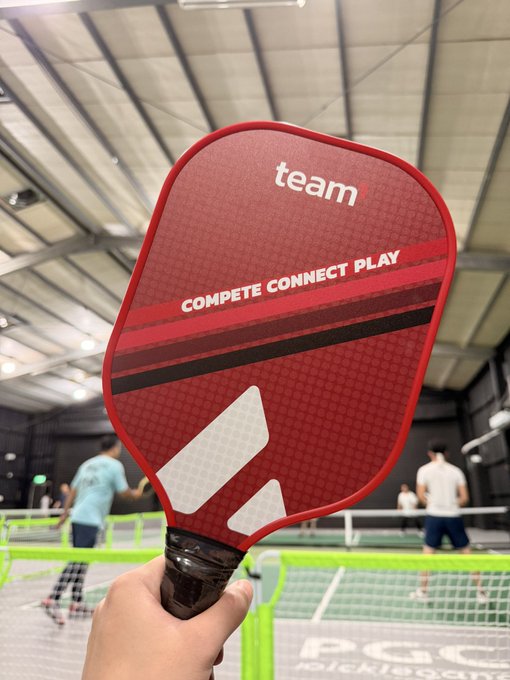 A hand holds up a red pickleball paddle with "team" branding and "COMPETE CONNECT PLAY" text, with a pickleball court visible in the background.