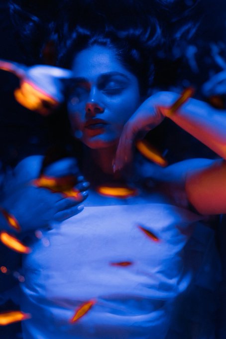 An atmospheric blue-tinted portrait photograph of a woman with closed eyes, surrounded by floating glowing embers or fireflies that create a dreamy, ethereal effect.