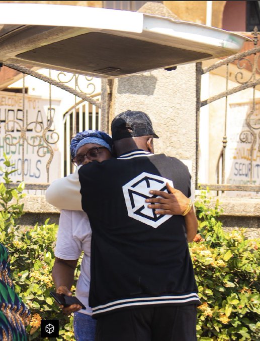 Two people share a heartfelt hug outdoors in front of a gated entrance with a partially visible "WELCOME TO" sign, while the man wears a black jacket featuring a geometric logo, symbolizing community and human connection that aligns with the post's message about blockchain gaming prioritizing player experience and seamless interaction over technical complexities.