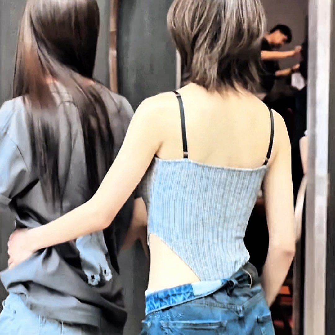 A back view of two women walking away, one in a black shirt and the other in a light blue ribbed tank top with thin straps and jeans, captured in a slightly blurred, casual setting.