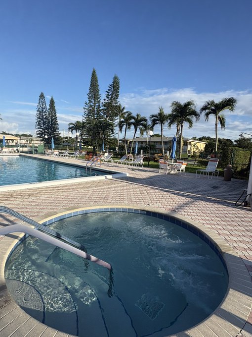 A resort pool area featuring a bubbling hot tub in the foreground, a larger swimming pool, lounge chairs, umbrellas, and palm trees under a partly cloudy blue sky.