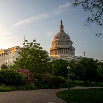 The United States Capitol building dome and grounds at golden hour, surrounded by green trees and a brick walkway.