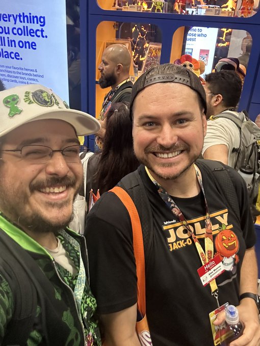 Two men smiling at what appears to be a convention, one wearing a cap with various pins and the other in a black "Jolly Jack-O" shirt with a conference badge.