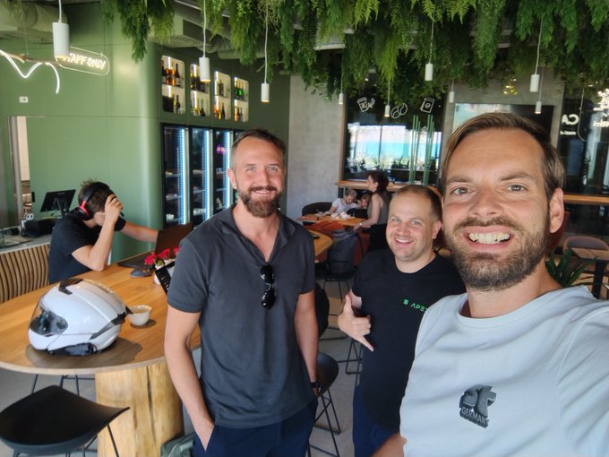 Three smiling men pose for a selfie in a cafe, with one wearing an 'APP' shirt and a motorcycle helmet resting on a nearby table.