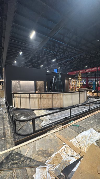 An interior view of a building under construction, showing a curved ride track or platform with metal railings, a trailing black backdrop, and ladders, with protective sheeting and lumber scattered on the floor.
