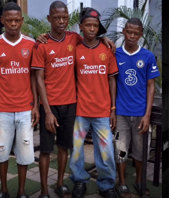 Four young men pose together outdoors wearing soccer jerseys: one Arsenal (red, 'Fly Emirates'), two Manchester United (red, 'TeamViewer'), and one Chelsea (blue, '3' sponsor).
