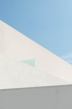 A minimalist photograph of a white architectural wall against a clear blue sky with a small triangular window.