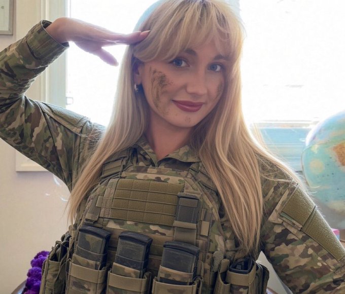 A woman in military camouflage uniform and tactical vest salutes, with dirt on her face and long blonde hair, posing indoors.