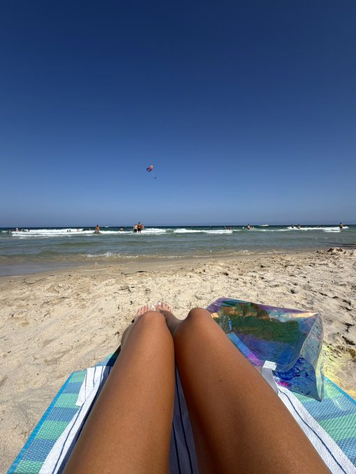 A first-person perspective from a striped beach towel showing a person's bare legs, with a clear blue ocean, sandy beach, and a kite in the sky in the background.
