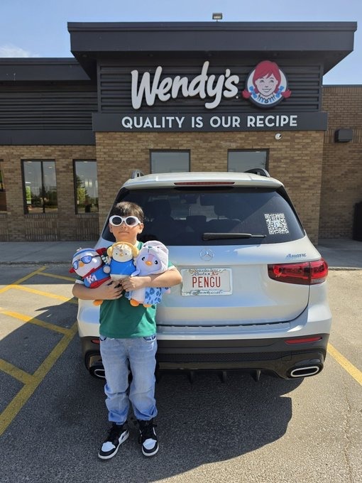 A young boy wearing sunglasses holding stuffed animals (including what appears to be the Wendy’s mascot) standing in front of a white Mercedes SUV with an Ohio Pengu license plate at a Wendy's restaurant.
