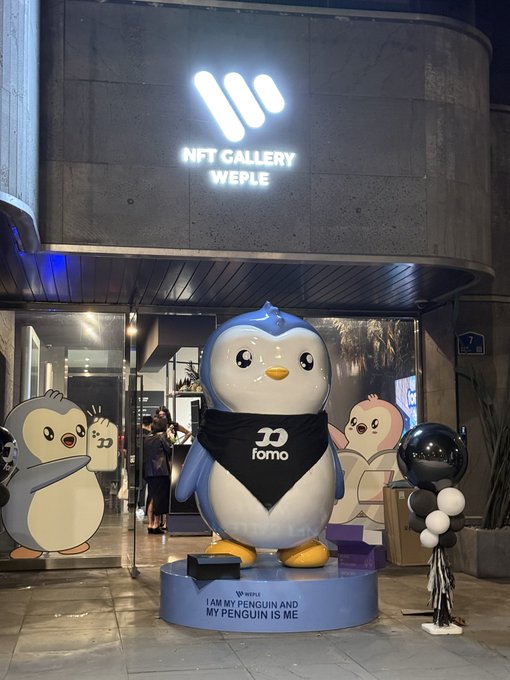 Nighttime photo of a large penguin statue wearing a "fomo" bandana outside an "NFT Gallery WEPLE" store, accompanied by smaller cartoon penguin cutouts and balloons.