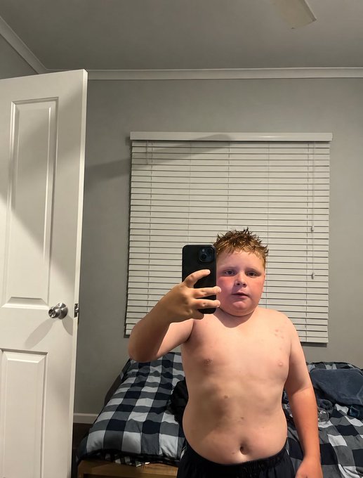 A shirtless young boy taking a mirror selfie with a smartphone in a bedroom, standing in front of a bed with a black and white checkered comforter.