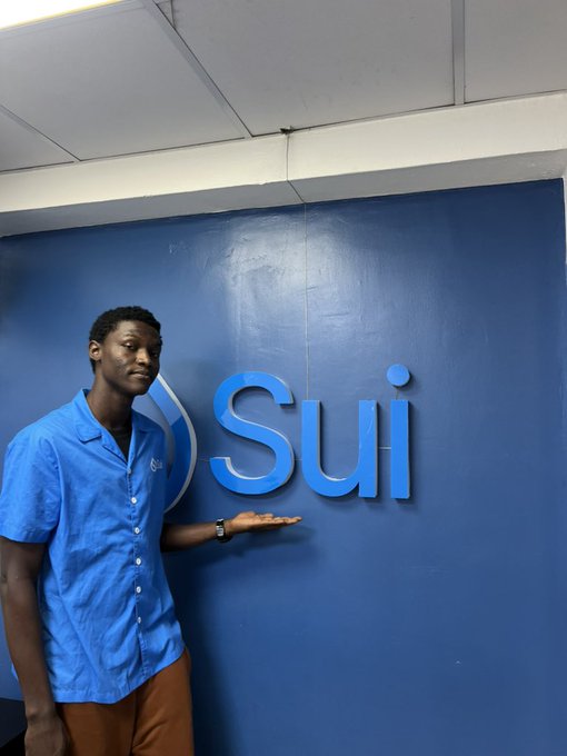 A man in a blue shirt stands against a blue wall displaying the 'Sui' logo, gesturing toward it with his hand.