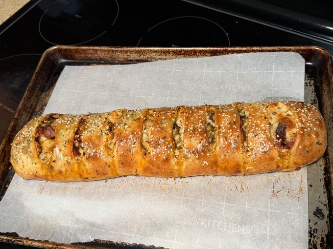 A freshly baked stuffed bread loaf topped with herbs and sesame seeds rests on a baking sheet lined with parchment paper on a stovetop.
