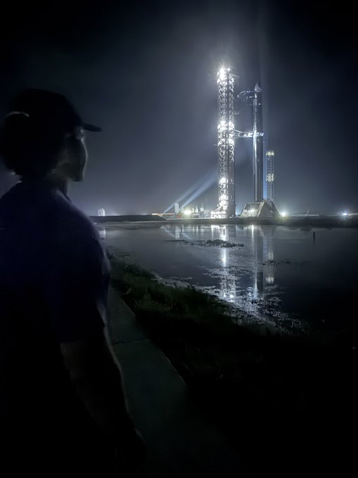 A person in silhouette watches a SpaceX rocket launch at night, with the brightly lit launch tower reflecting in the water below.