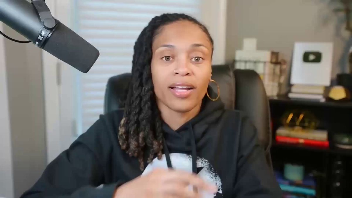 The image shows a woman sitting behind a microphone, possibly giving a speech or participating in an interview. She has long dreadlocks and is wearing a black hoodie. The background of the image features a bookshelf filled with various books, suggesting that she might be discussing topics related to literature or education.