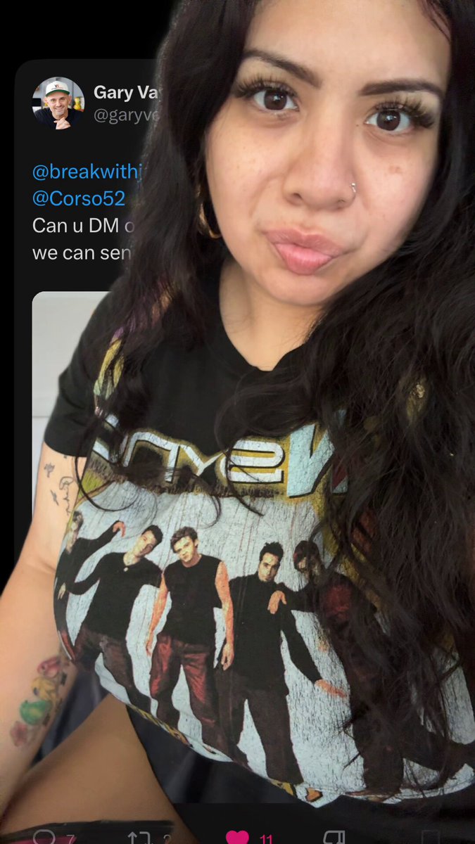 The image features an attractive young woman who has a tattoo on her arm, wearing a black t-shirt with various band logos printed across the front. She appears to be taking a selfie using her phone while sitting or standing. The caption of the image is related to the posts, as it mentions that she just received a comic in the mail and will be a Vee Friends holder by the end of 2026.