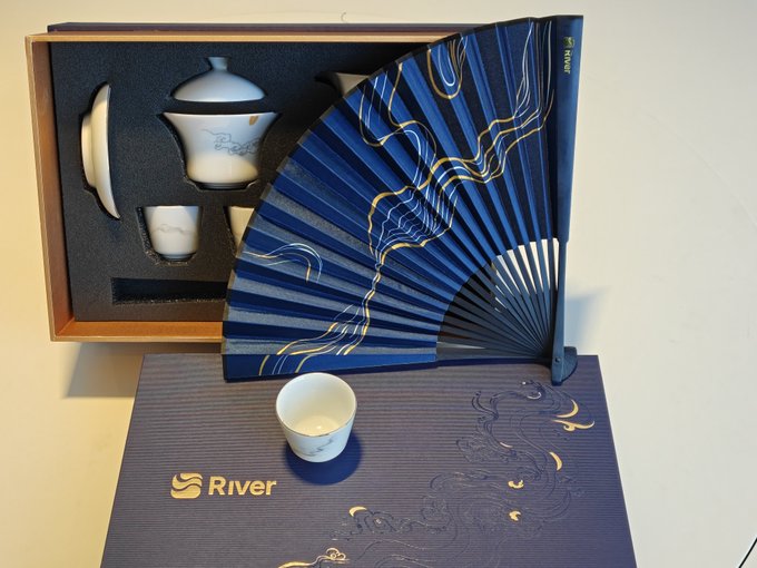 An elegant gift set from "River" featuring a traditional Chinese tea set (gaiwan and cups) displayed alongside a decorative blue and gold folding fan and a patterned box, resting on a dark mat with golden dragon designs.